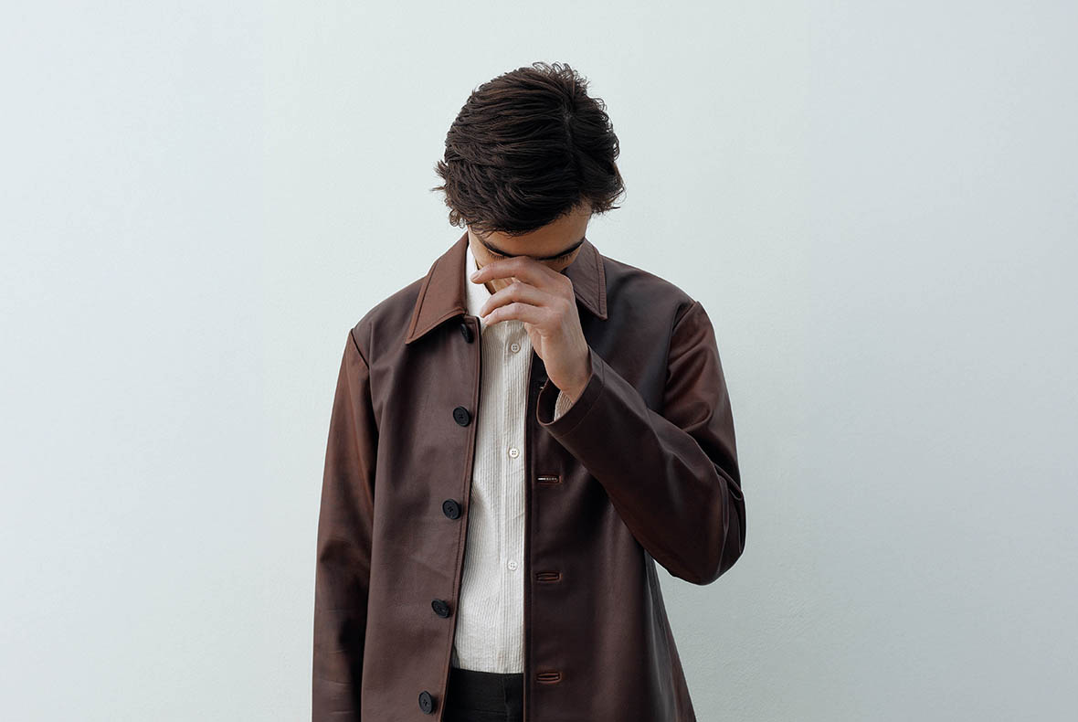 Young man wearing a brown coat and white shirt, looking down with hand near his face against a pale grey background