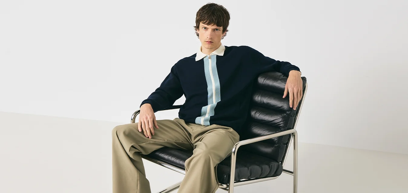 Giovane uomo in maglione blu navy con striscia verticale e pantaloni beige seduto su una sedia in pelle nera e metallo, contro uno sfondo bianco.
