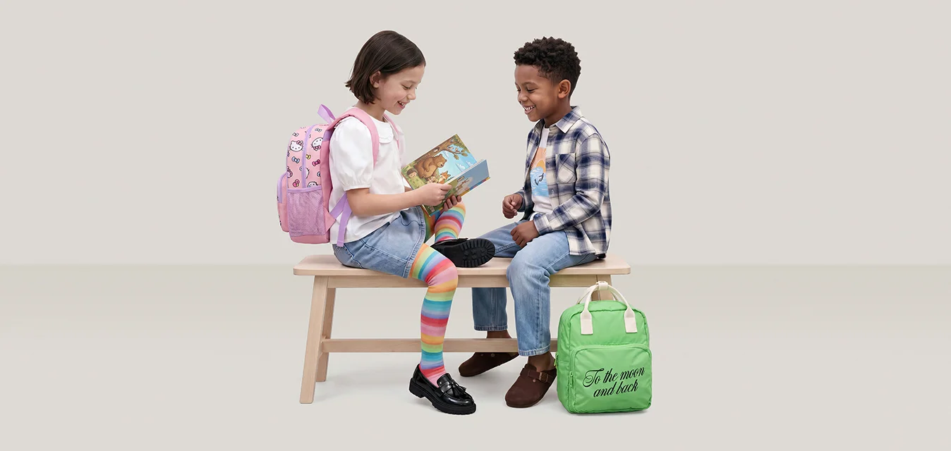 Due bambini sono seduti su una panchina di legno; uno legge un libro mentre l’altro ascolta, circondati da zaini colorati.