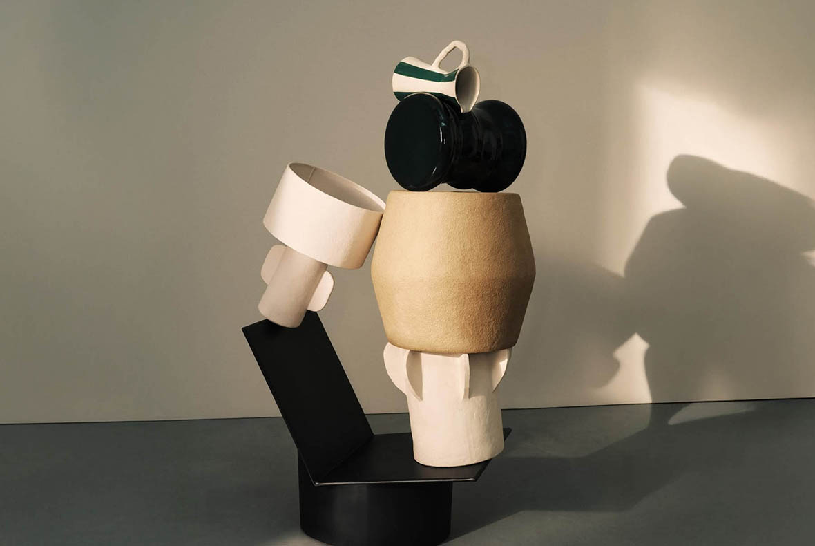 Abstract stacked ceramic sculpture of beige, black and white vessels with a green-striped jug on a pedestal casting a shadow