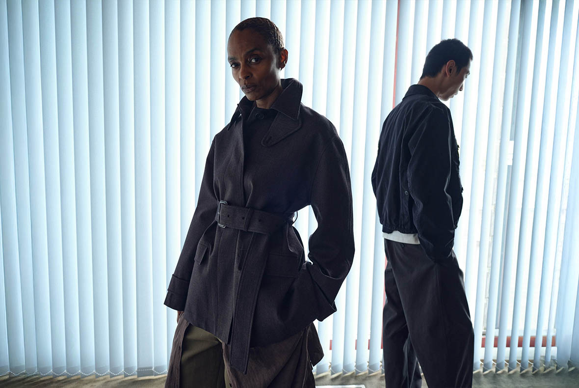 Two fashion models by vertical window blinds, foreground model in belted dark trench coat, moody backlit studio portrait