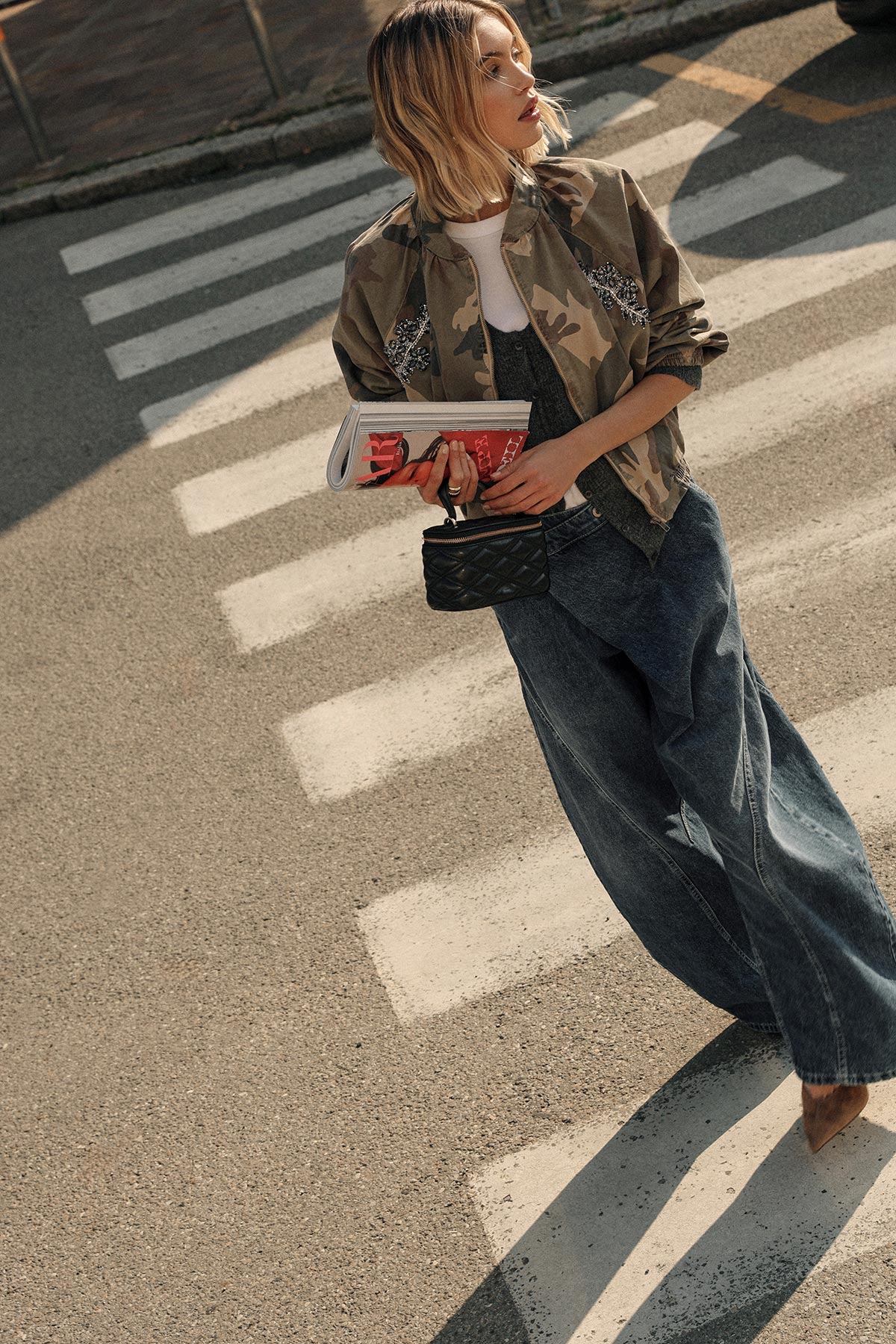 Woman crossing zebra crossing wearing camouflage jacket and wide jeans, holding magazines and a quilted black handbag, looking sideways
