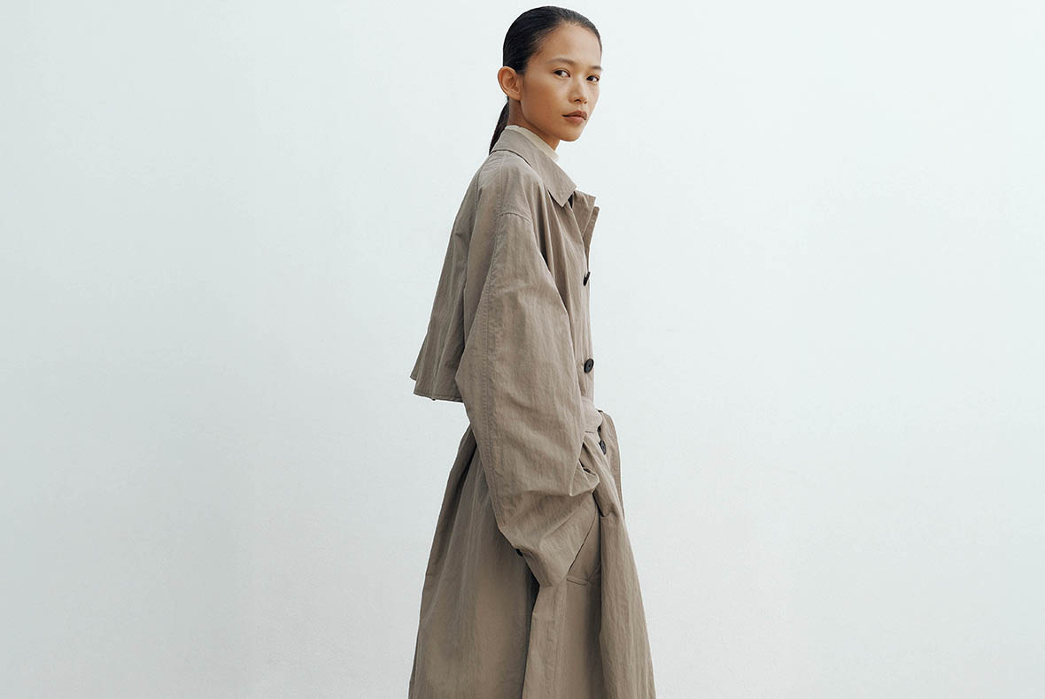Female model wearing a beige oversized trench coat, standing sideways with hands in pockets against a pale neutral background