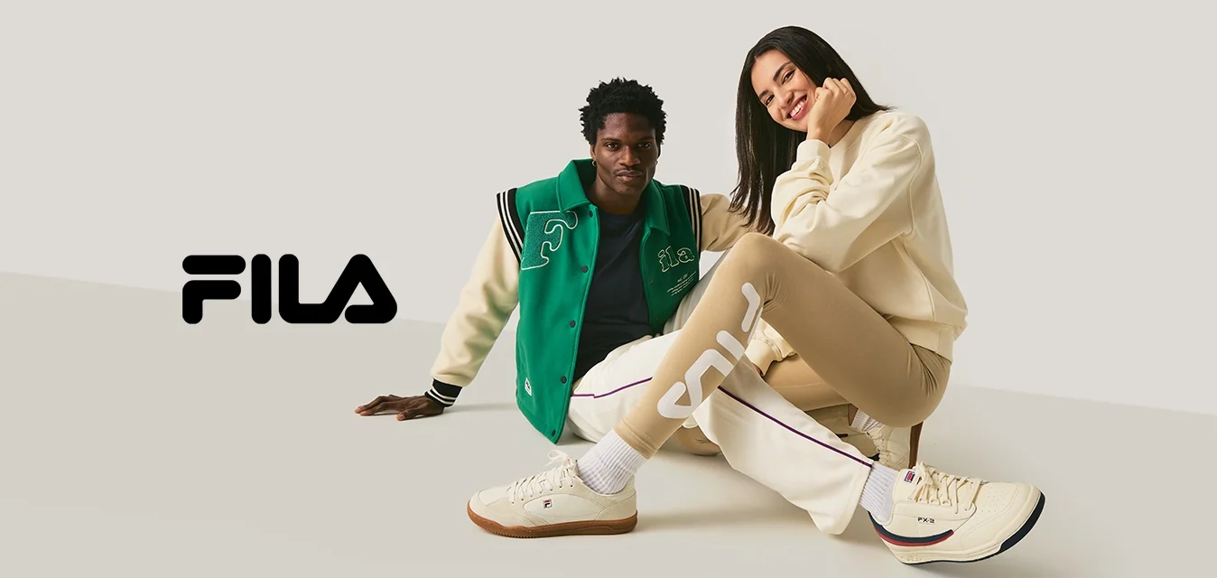 Due persone sedute per terra, una indossa una giacca varsity verde e bianca, l'altra dei leggings beige e una felpa crema, sorridenti.