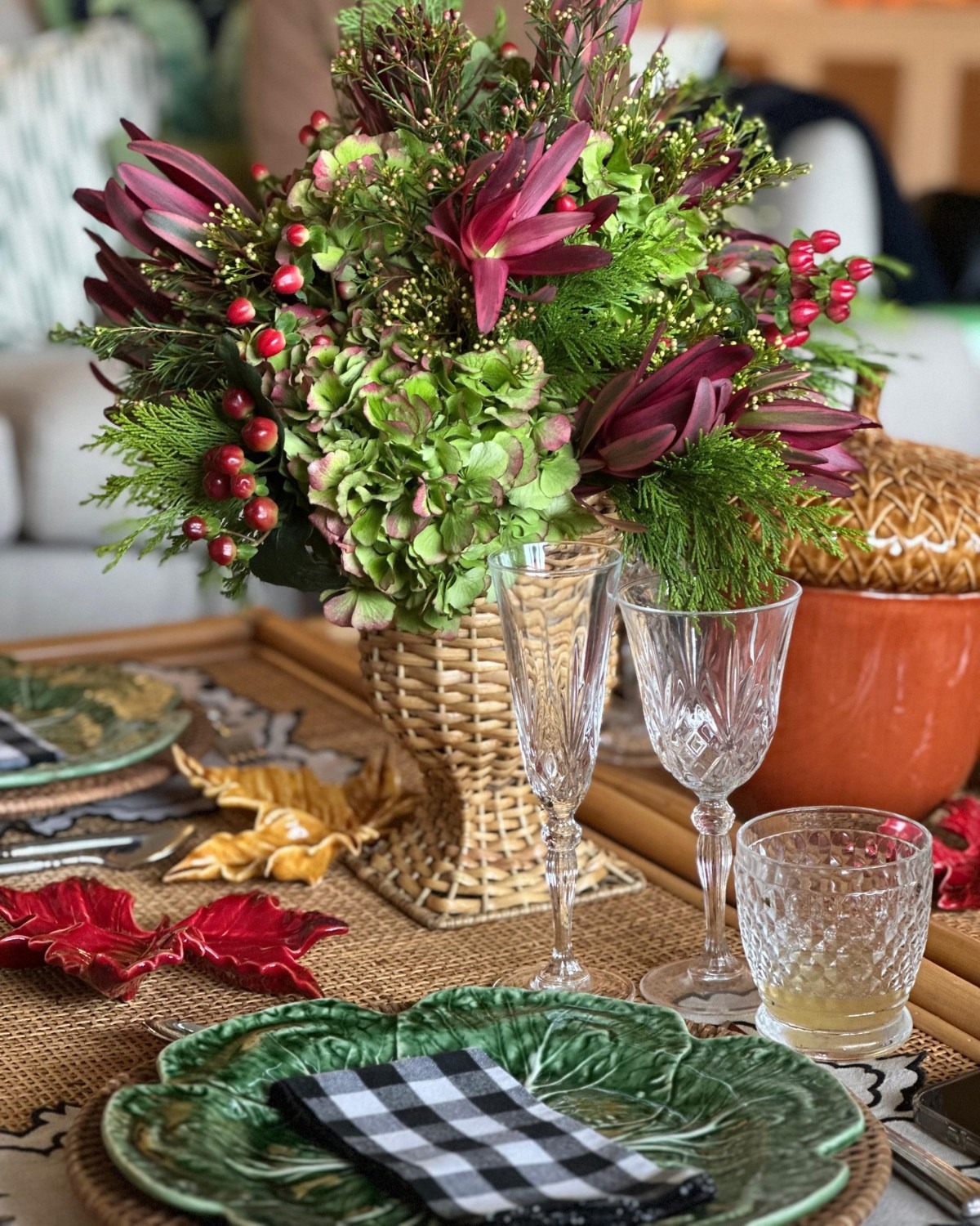 Festive table setting with wicker vase of green hydrangea, burgundy lilies and red berries, crystal glasses and green leaf plate with checkered napkin