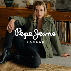 Donna bionda con giacca verde, jeans neri e stivali, seduta a terra appoggiata a un mobile in legno con dischi in vinile e vasi decorativi.