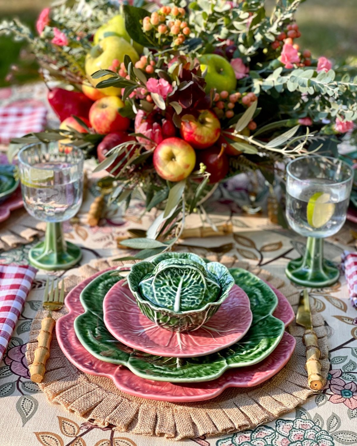 Cottage-style outdoor table setting with layered green and pink cabbage-shaped ceramic plates, floral and apple centrepiece and water glasses