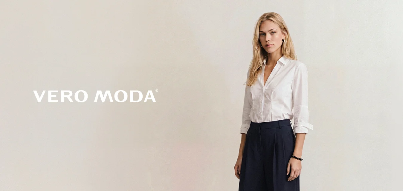 Donna in piedi che indossa una camicia bianca abbottonata, pantaloni neri a gamba larga, scarpe nere e un bracciale nero su uno sfondo semplice.