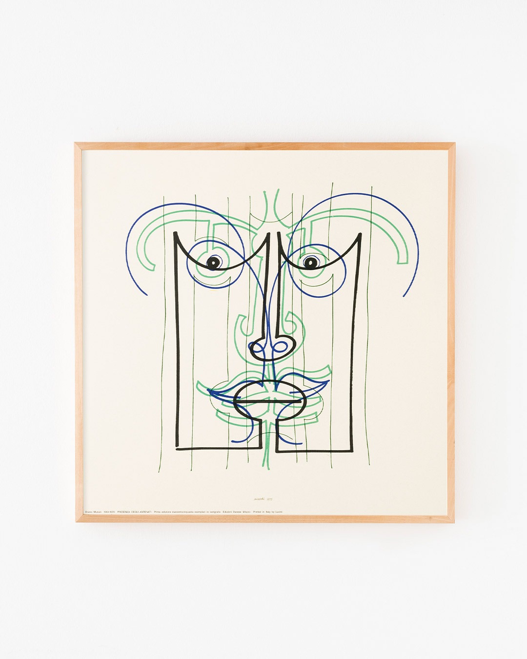 Framed abstract line-art face print in black, blue and green on cream paper with a thin wooden frame mounted on a white wall