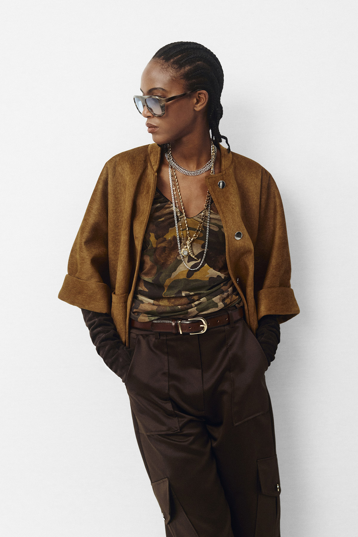 Stylish Black woman in sunglasses wearing a brown suede jacket, camouflage top, layered chains and high-waist cargo trousers, hands in pockets