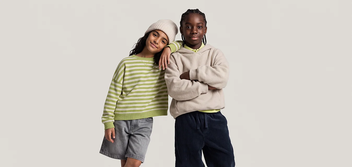 Due bambini in piedi uno accanto all'altro, uno appoggiato sulla spalla dell'altro, vestiti con abbigliamento invernale casual su uno sfondo semplice.