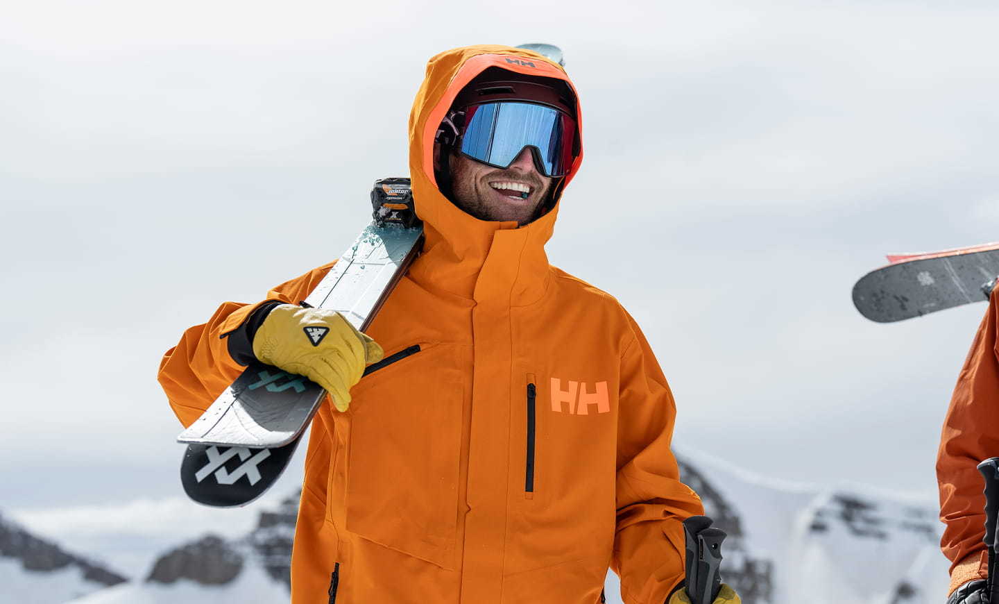 Man wearing an orange Helly Hansen ski jacket, carrying skis on a snowy mountain.