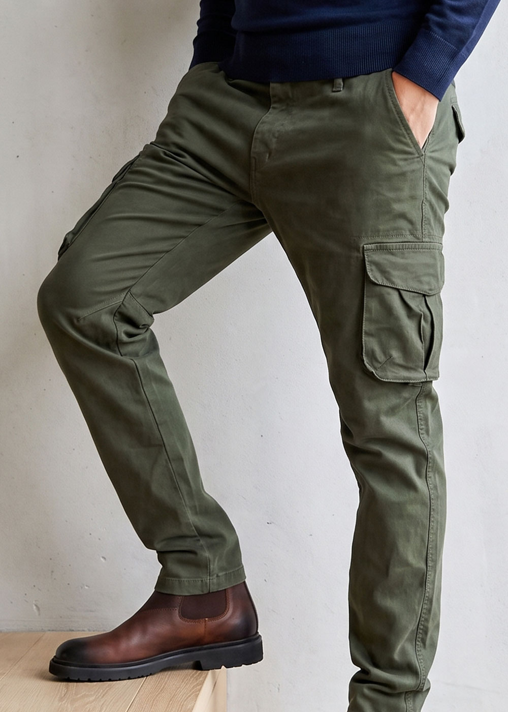 Man wearing olive green cargo trousers and brown leather Chelsea boots, hands in pockets with navy jumper visible