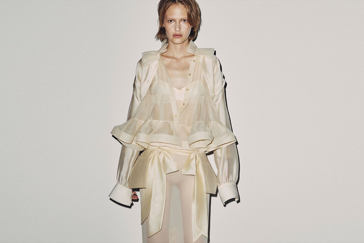 Female model wearing cream sheer ruffled blouse with oversized sleeves and tied satin bow belt, standing against pale backdrop