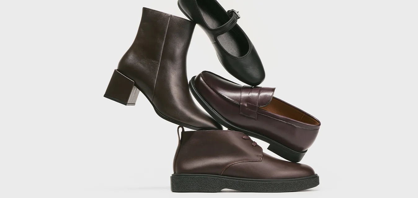 Scarpe impilate di colore marrone e nero, tra cui un stivale con tacco, una Mary Jane, un mocassino e una scarpa casual allacciata, su uno sfondo grigio chiaro.