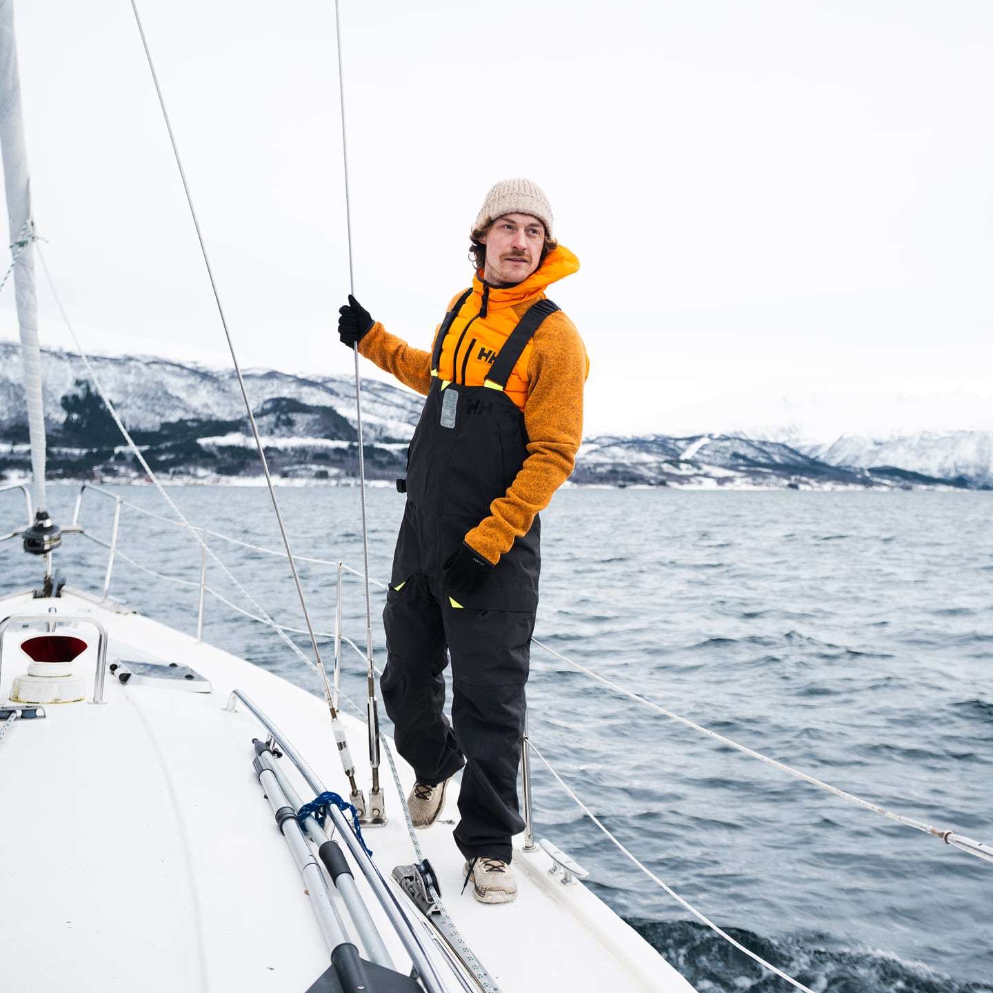 Man wearing an orange Helly Hansen jacket and black sailing bib overalls on a boat in cold waters.