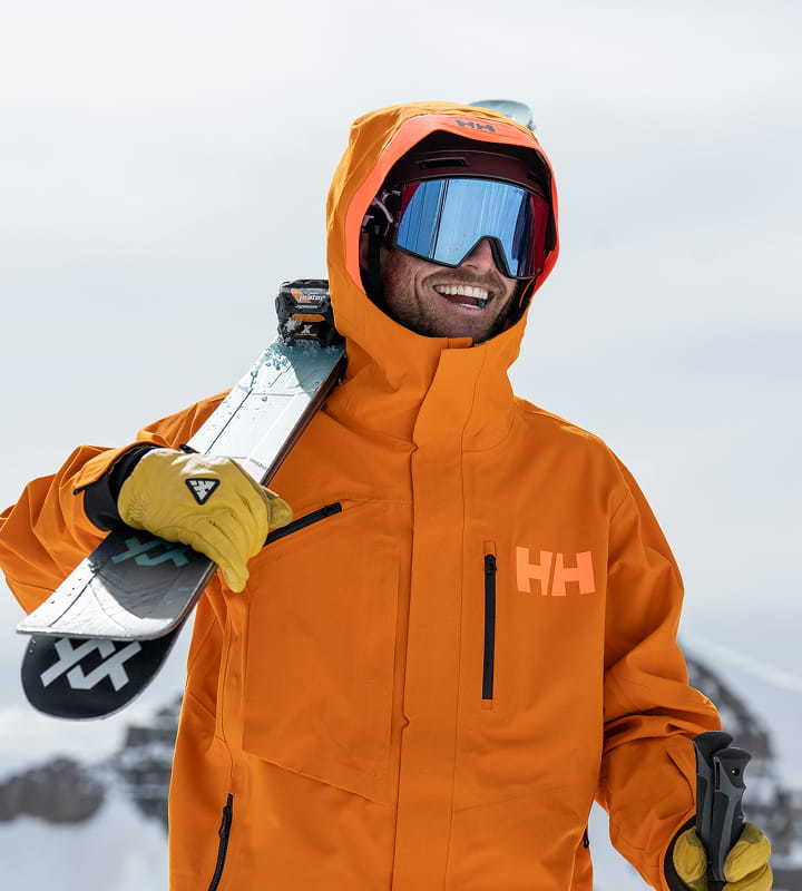 Man wearing an orange Helly Hansen ski jacket, carrying skis on a snowy mountain.