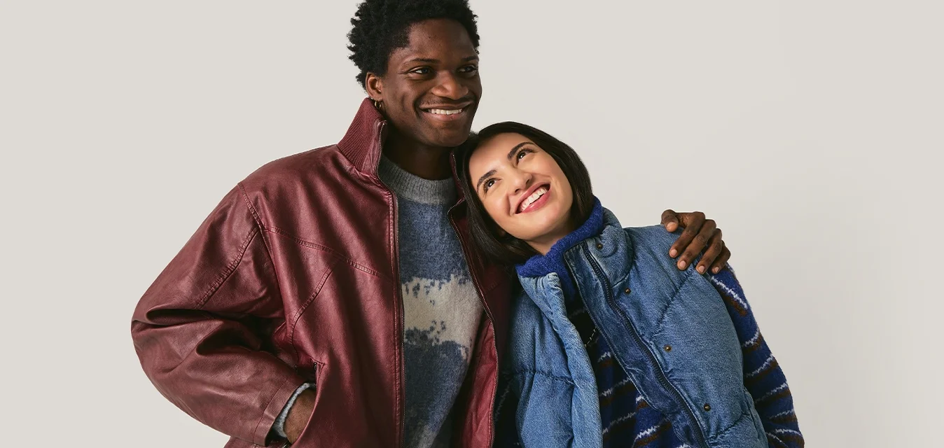 Uomo sorridente con un braccio intorno alla donna sorridente che appoggia la testa sulla sua spalla, entrambi indossano giacche casual su uno sfondo chiaro uniforme.
