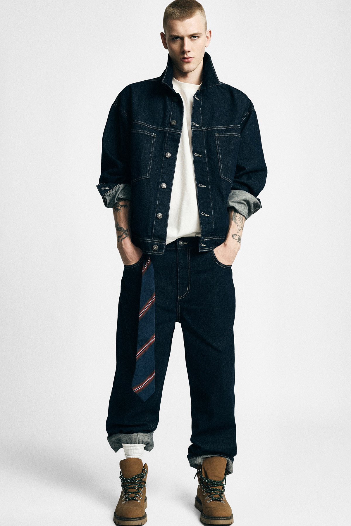 Young man model in dark denim jacket and jeans, white T‑shirt, striped tie hanging from belt, rolled cuffs and tan boots, visible tattoos