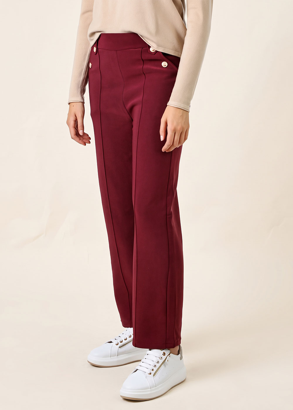 Woman wearing burgundy tailored high-waist trousers with gold buttons, beige top and white trainers, side view