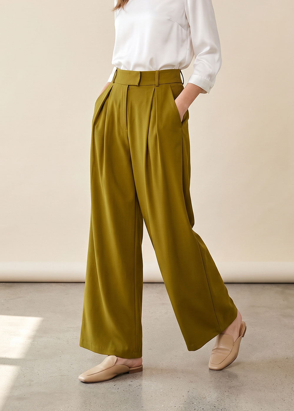 Model wearing olive-green wide-leg pleated trousers with white blouse tucked in, hands in pockets and beige loafers