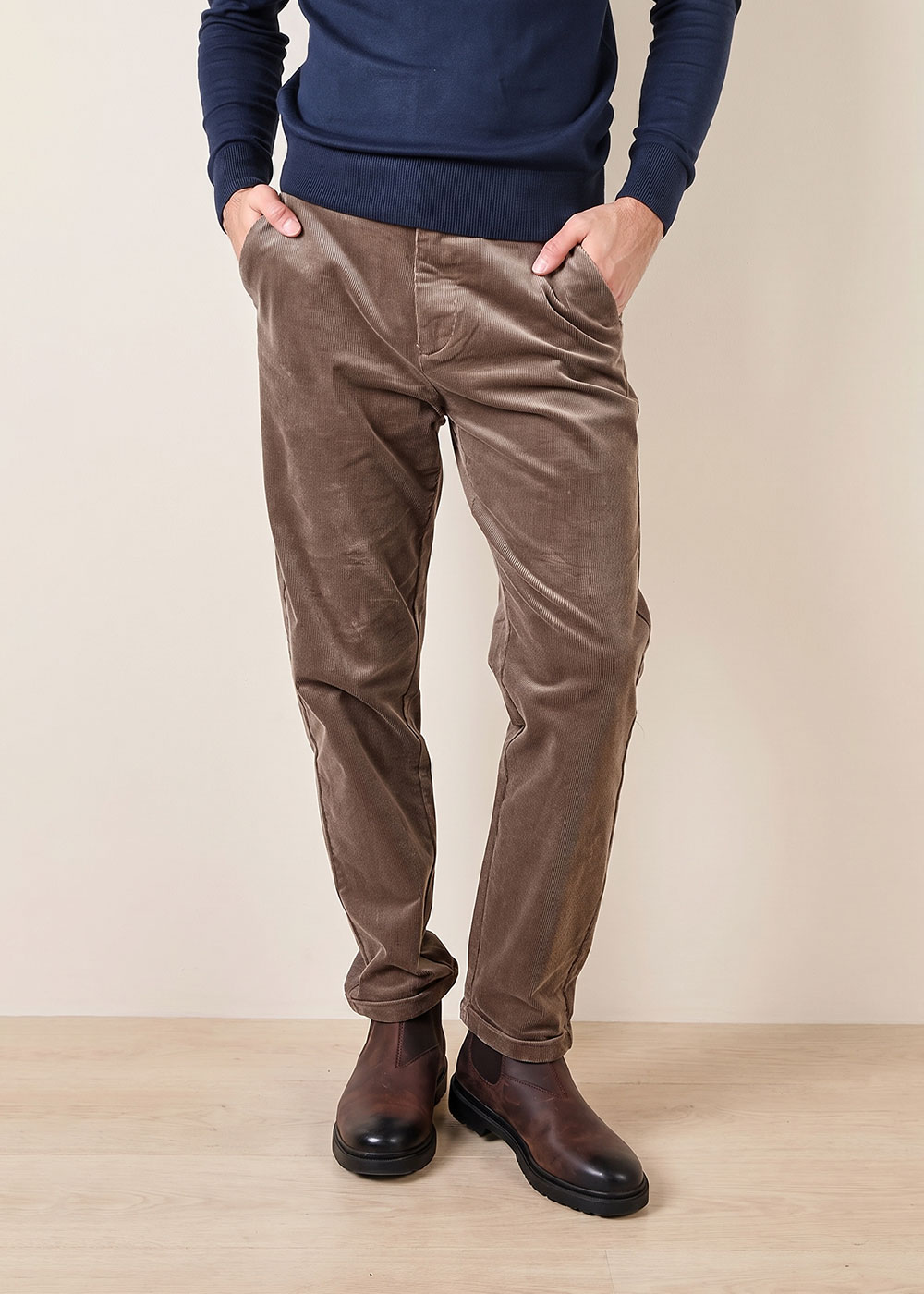 Man wearing brown corduroy trousers and brown leather Chelsea boots with navy jumper, hands in pockets, standing on floor
