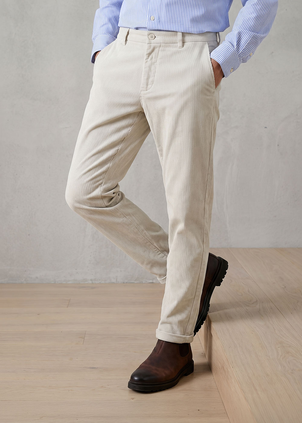 Man wearing beige corduroy trousers with a blue striped shirt tucked in and brown leather Chelsea boots in a relaxed leaning pose