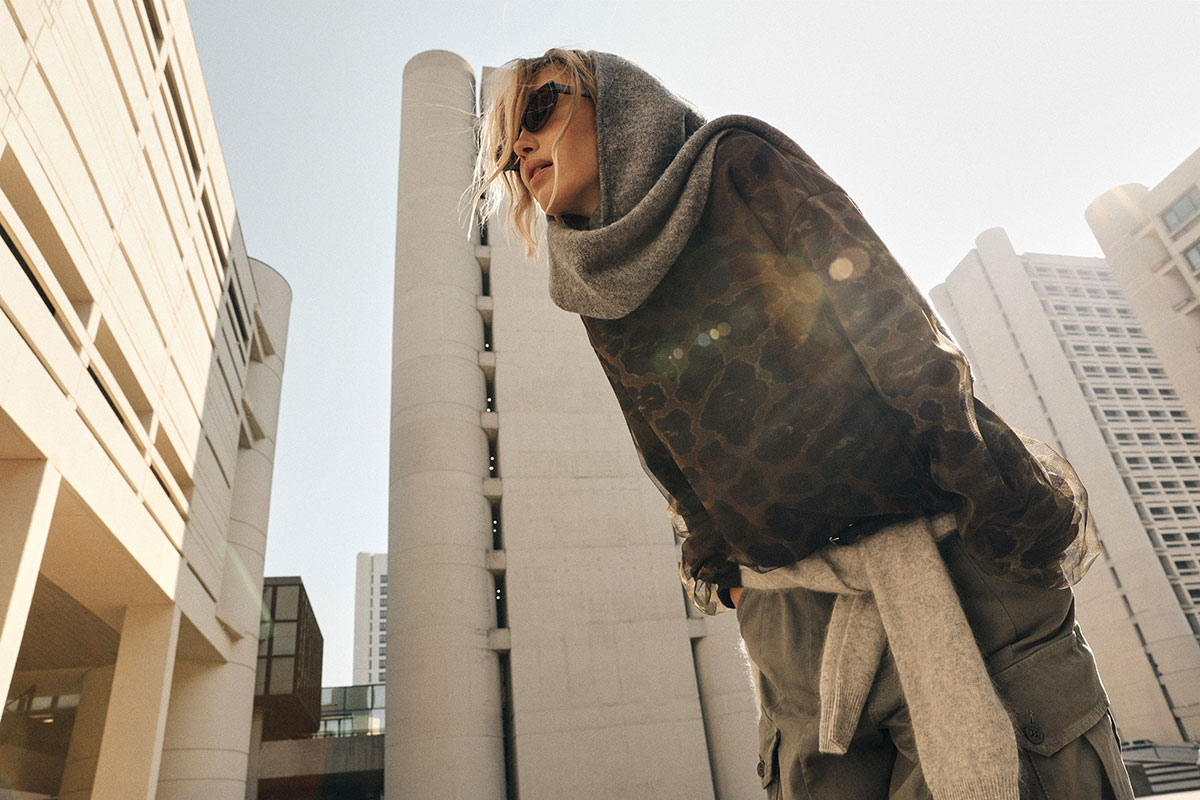 Stylish woman in sunglasses and grey hooded scarf leaning forward in a modern urban plaza with sunlight flare