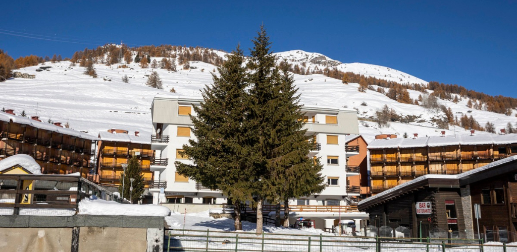 Snow-covered alpine resort with multi-storey chalets, large central fir tree and sunlit mountain slope under a blue sky