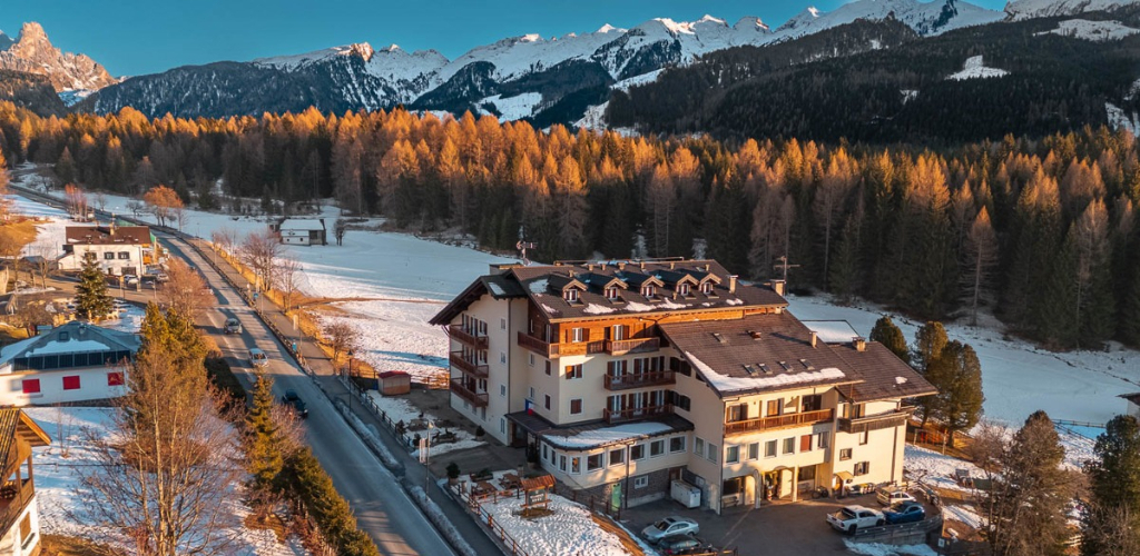 Alpine hotel and chalets beside a snowy road, sunlit pine forest and snow-capped mountains at golden hour