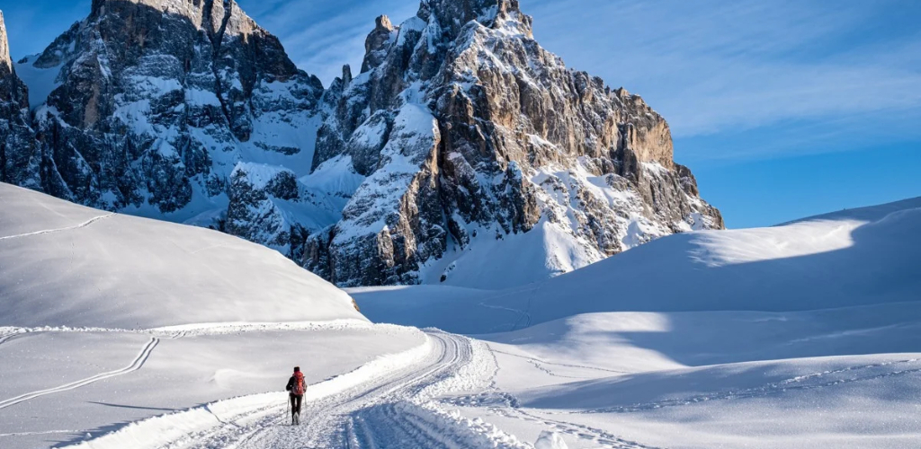 Lone hiker on snowy trail with ski tracks leading towards rugged snow-covered mountain under clear blue sky