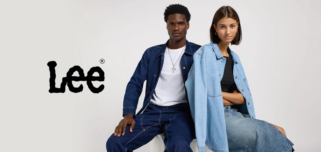 Uomo e donna seduti fianco a fianco su un blocco bianco, entrambi vestiti con abiti di denim su uno sfondo chiaro e uniforme.