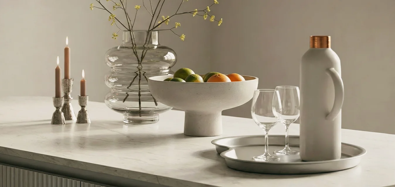Isola cucina in marmo con vaso di vetro con fiori gialli, ciotola di frutta, tre candelabri e vassoio con brocca bianca e due bicchieri da vino.