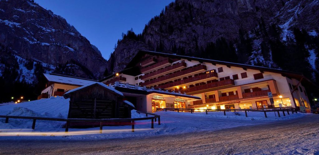 Snow-covered alpine hotel and chalet at dusk with warm glowing windows, illuminated mountain valley road
