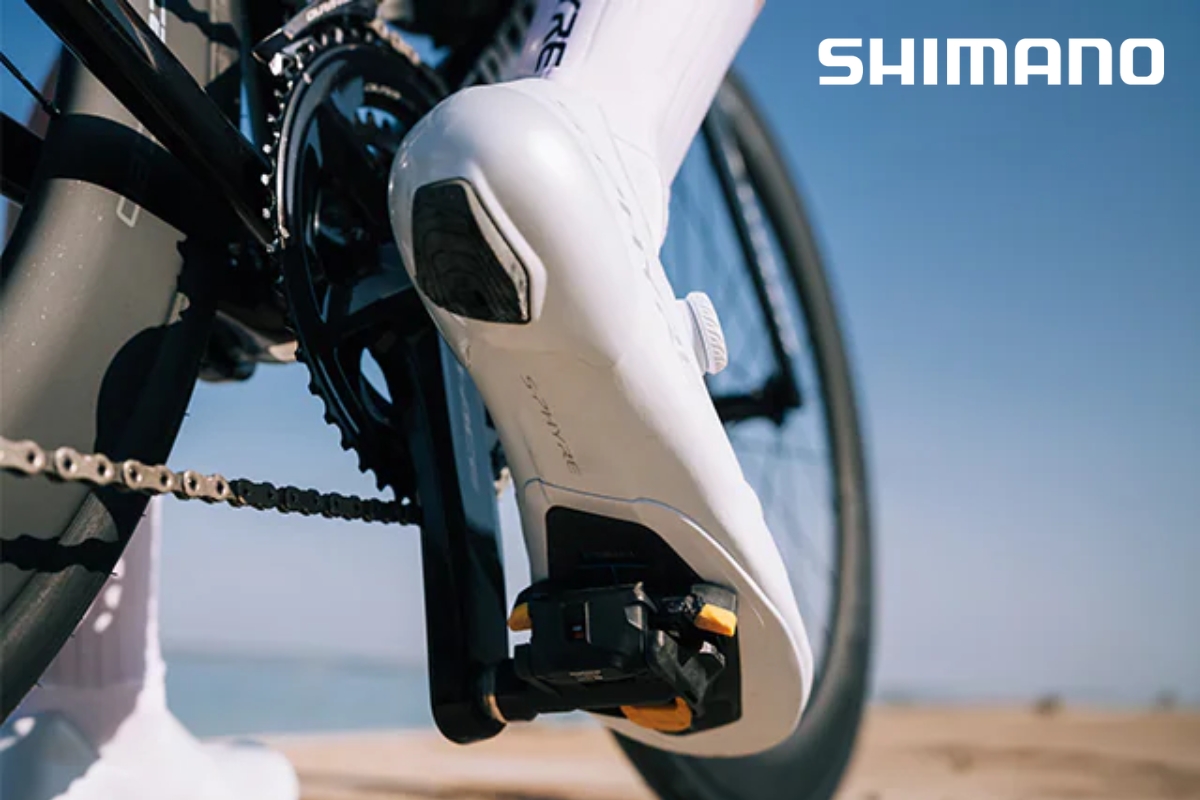 Close-up of white cycling shoe clipped into a Shimano clipless pedal on a road bike with beach and blue sky background
