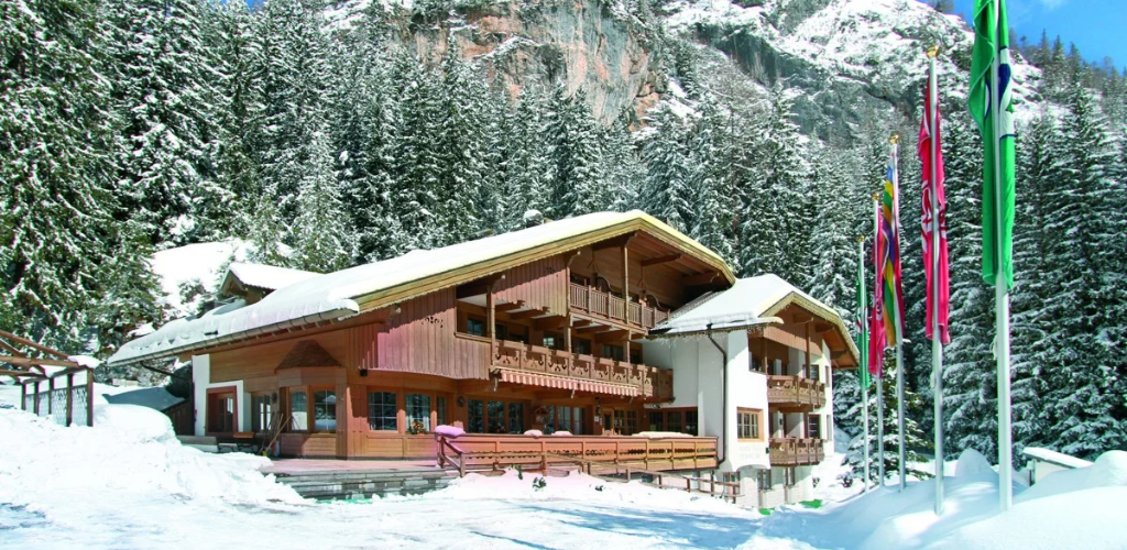 Snow-covered wooden alpine chalet with balconies beside snow-laden pine forest and colourful flags on a mountain backdrop