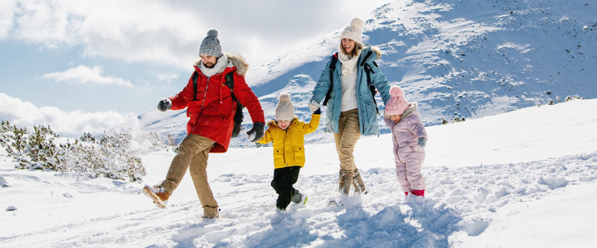 Family of four (two adults, two children) walking hand‑in‑hand on a snowy mountain slope, wearing colourful winter coats and hats