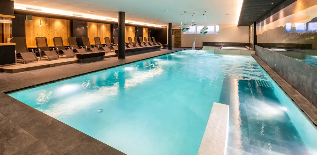 Indoor luxury hotel pool with turquoise water, submerged benches, wellness jets and row of reclining loungers in modern spa.