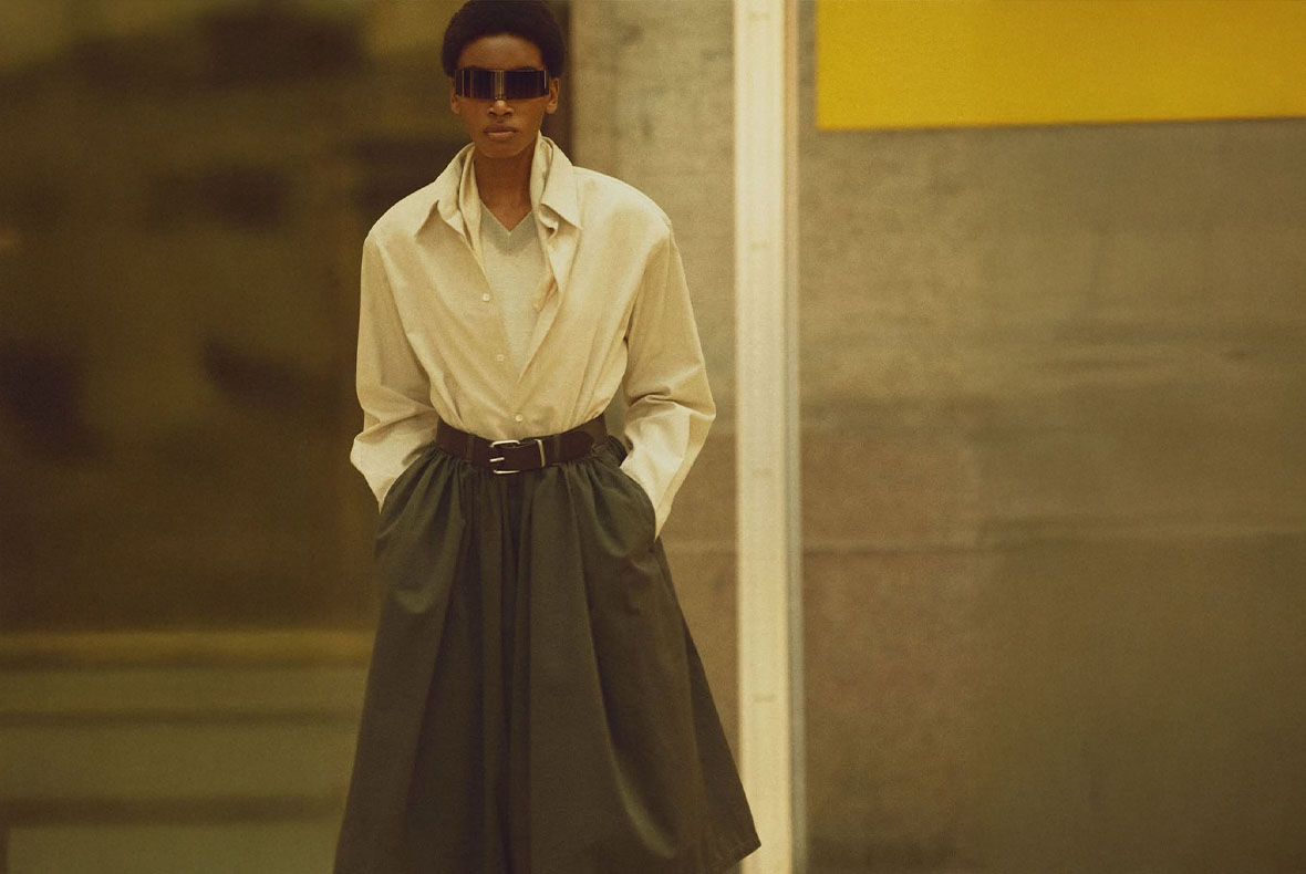 Fashion model in oversized cream shirt, cinched dark full skirt and futuristic visor sunglasses standing against urban wall