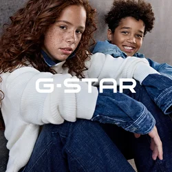 Due bambini, uno con capelli rossi e ricci e un maglione bianco, e uno con capelli ricci corti con una giacca di jeans, seduti contro un muro di cemento.