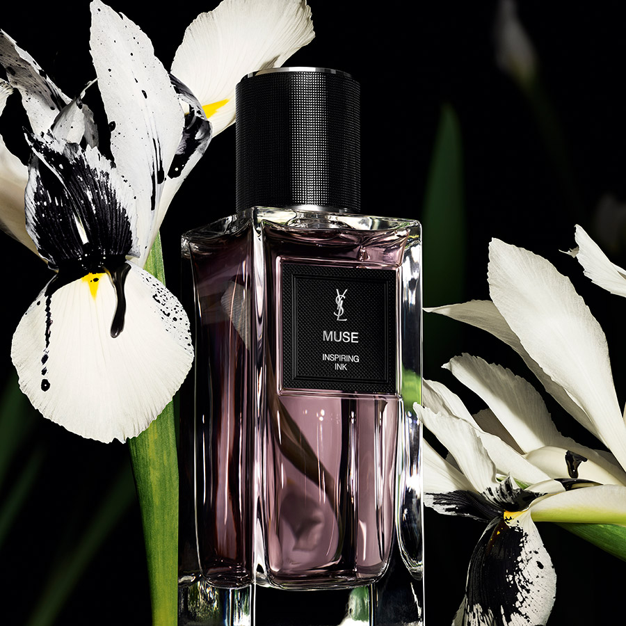 Un flacone di profumo YSL Muse circondato da iris bianchi con schizzi d'inchiostro nero.