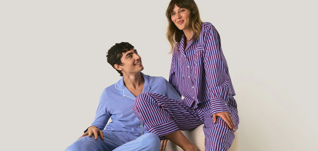 Due persone vestite con pigiami a righe sedute e in piedi su un sedile imbottito beige contro uno sfondo semplice.