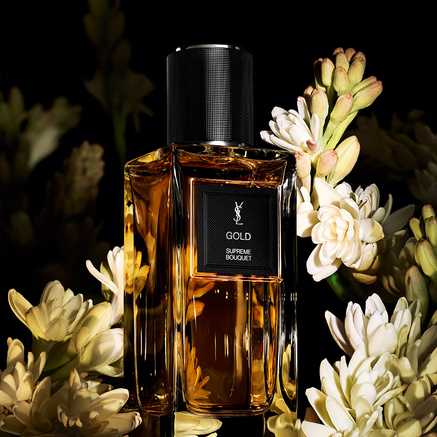 Un flacone di profumo YSL Gold Supreme Bouquet circondato da fiori di tuberosa bianchi su sfondo scuro.