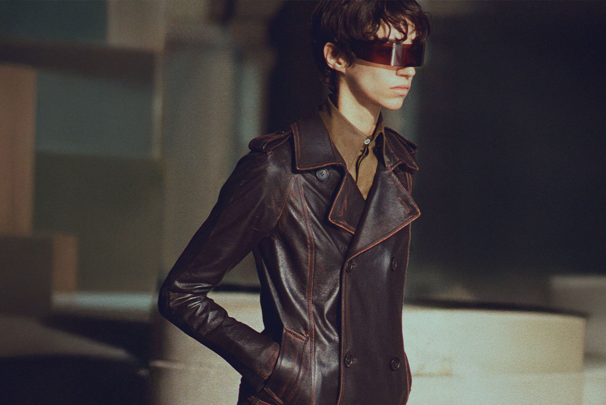 Model wearing dark brown leather trench coat and futuristic visor sunglasses, hands in pockets, moody studio lighting