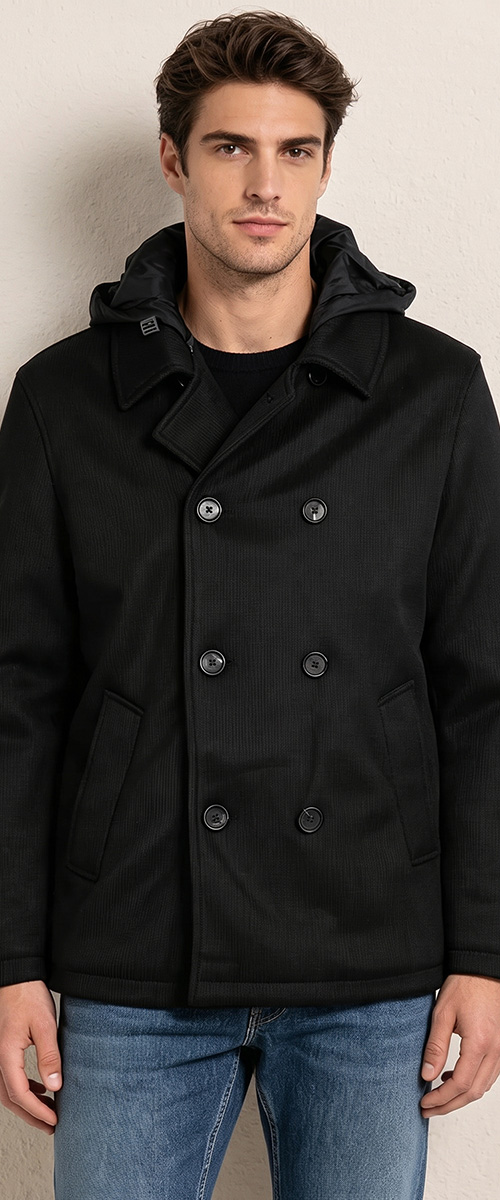 Male model wearing a black double-breasted hooded pea coat and blue jeans against a beige wall