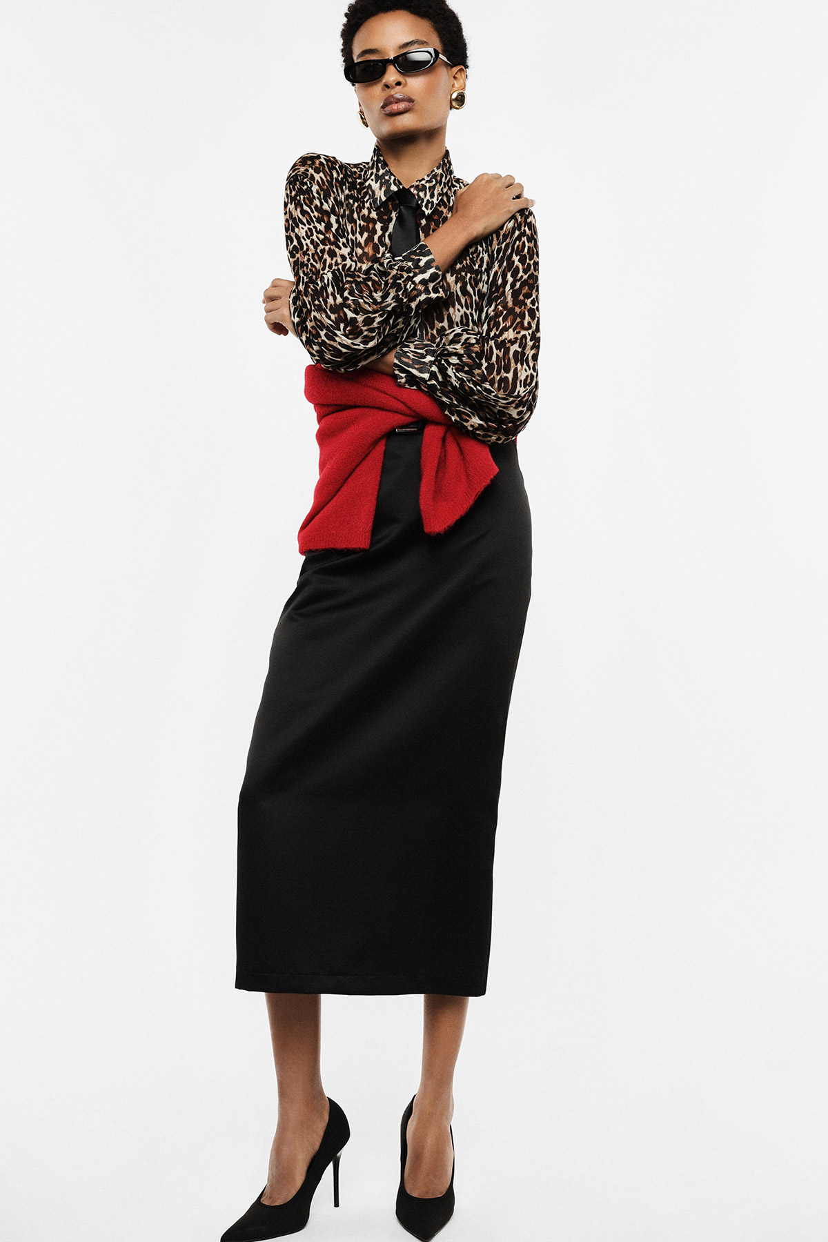 Woman wearing leopard-print blouse, black midi skirt, red sweater tied at waist, black heels and sunglasses, arms crossed