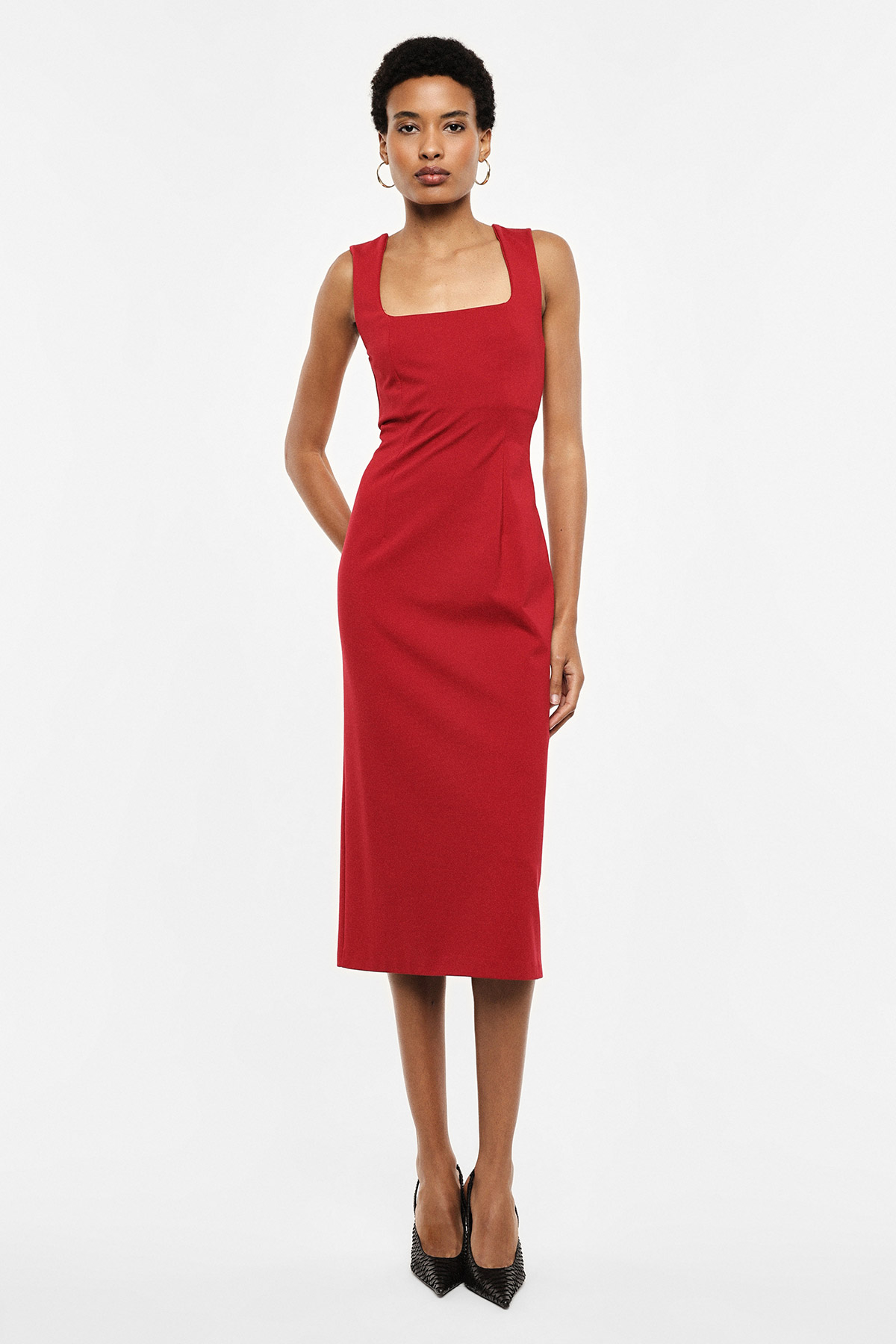 Woman in a red square‑neck sleeveless midi dress with hoop earrings and black pointed heels on a white background