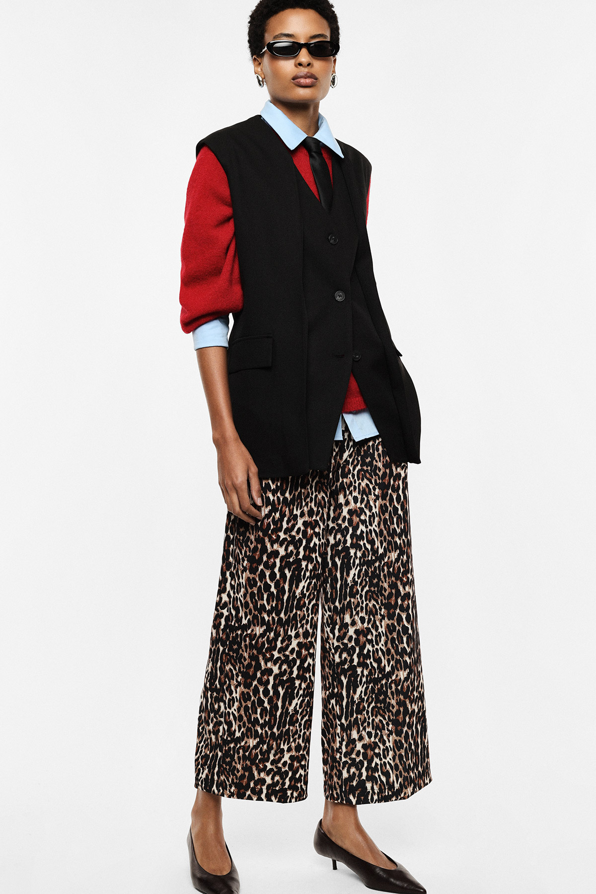 Model wearing black sleeveless blazer over red sweater and blue shirt, leopard-print wide-leg trousers and black kitten-heel pumps