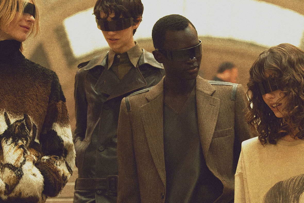 Four fashion models in muted brown tones wearing futuristic blindfold-style visors and tailored jackets in an indoor setting