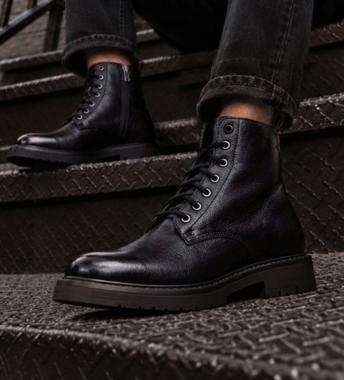 Close-up of black leather lace-up ankle boots on textured metal stairs, rugged sole and eyelets, urban streetwear style